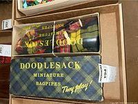 Photo showing the original Doodlesack box with the miniature bagpipes inside, clear window on top, and tartan plaid pattern on box.