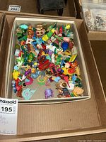 Cardboard box filled with assorted small plastic toys of various shapes and colors, including vehicles and animal figures