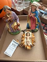 Three nativity figurines in a box: Joseph, Mary, and baby Jesus in a golden manger.