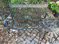 Front view showing black metal outdoor bench with lattice seat and decorative backrest on brick patio with leaf litter and garden in background. Some rust and corrosion visible on seat base.