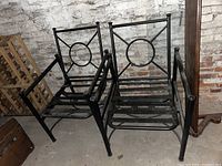 Two black metal outdoor patio chairs placed next to a wall, showing the front and side views. Chairs have a circular and X pattern on the backrest and slatted seat base.