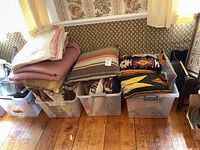 Photo showing multiple decorative pillows and folded blankets stacked on plastic bins on wooden floor.