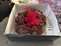 Pinecone wreath inside a white box placed on a carpet, showing the wreath decorated with different pinecones and a central red bow. A MaxSold sticker (2x4 inch) is visible for scale.
