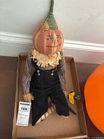 Full view of the pumpkin head scarecrow doll sitting in a box, showing outfit and overall condition.