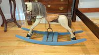 Side view of wooden rocking horse showing cream body, multi-colored mane and tail, brown leather saddle with studs, and blue rocking base.