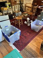 Wide view of the red patterned area rug with wooden floor visible around it and various items such as chairs, plastic bins, and small stools on and around it.