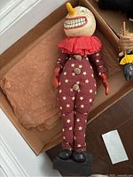 Front view of full clown doll in box showing large round head with painted face, red ruffled collar, star-patterned cloth body, red gloves, and black shoes on stand.
