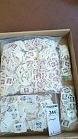 Cardboard box filled with numerous used USA postage stamps on paper backing, arranged in loose piles and small bags.