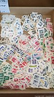 Cardboard tray containing scattered used U.S. postage stamps affixed to paper backing, featuring various colors and printed designs.
