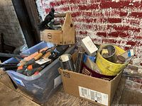 View of assorted hand and garden tools in plastic tub and cardboard box, wooden toolbox, and various small hardware items all placed on floor against a brick wall.