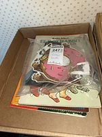 Photo of vintage items in cardboard box, showing a bagged set of monster masks including a pink donut-shaped mask, and vintage books below.
