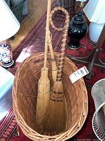 Photo showing two vintage brooms in a wooden basket, placed on a patterned carpet with nearby furniture.