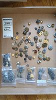 Top-down view of a cardboard display board featuring a scattered assortment of loose vintage buttons and small plastic bags with more buttons.