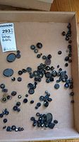 Top-down view of many vintage black buttons scattered in a cardboard box, showing varied sizes and shapes.