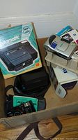 Box containing vintage cameras including an old Kodak Kodamatic instant film camera in its original packaging, a silver camera, batteries, and manuals.