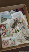 Photo of a box with assorted vintage advertising and collectible cards showing children's cartoons, floral art, and Victorian styled portraits.