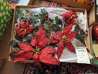 Two boxes containing vintage Christmas decor with large red poinsettia flowers, green holly ivy leaves, red berry clusters, and curled twine accents