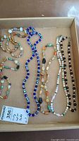 Five vintage costume jewelry necklaces on display in a cardboard box; featuring various colors and types of beads including stones and glass beads in natural and vibrant tones.