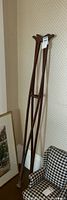 Photo of two vintage wooden crutches standing against wall next to patterned armchair and framed pictures on floor.