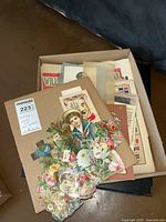 Photo showing the box containing vintage greeting cards and ephemera with scrap and die-cut floral designs and a sailor boy figure.