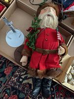 Full view of handcrafted vintage Santa Claus doll lying down in box with metal stand next to it.