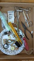 Photo showing an assortment of vintage household tools including scissors, a small hammer, screwdriver, keys, a magnifying glass, and a small clock face, all arranged in a cardboard box with a floral tray.