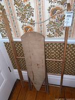 Vintage wooden ironing board with fabric padded top leaning against a wall with patterned wallpaper