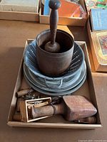 View of wooden mortar and pestle inside metal colander along with stacked pie plates and various small items including cock screws and clothes pins