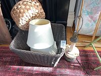 Photo showing the lamp base, large dark wicker basket containing two white lamp shades, wooden basket, and smaller wicker basket on a patterned carpet.