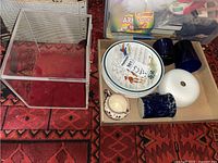 Photo showing vintage plates, ceramic mug, creamer, blue glasses, white vase or urn on a red patterned carpet and nearby cardboard box.