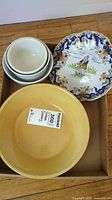 Top view of all bowls in a box showing white ironstone bowls stacked, large yellow pottery bowl, and decorative scalloped-edge pottery bowl with a figure and floral design