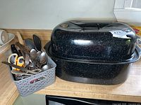 Photo showing vintage black enamel roasting pan with white speckles, next to gray plastic basket containing assorted kitchen utensils including scissors, flatware, spatula, and spoons.