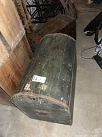Photo showing large dark green vintage wooden trunk with curved top, metal handles and studs, placed in basement with other wooden furniture around