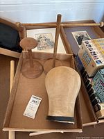 Top-down photo of the wooden spindle, two wooden wig holders/stands, and fabric-covered wig head block arranged inside a cardboard box alongside boxed puzzles.