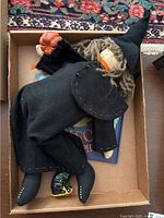 Top view of the witch doll lying in an open box showing the black dress, cape with stitched edges, pointed hat, yarn hair, and the pumpkin accessory. The small black glittery cat figurine is visible at the foot of the doll along with part of the book cover.