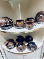 Photo of multiple brown ceramic cups and saucers with textured pattern displayed on a curved glass shelf.