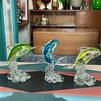 Front view of three art glass dolphins, two green and one blue, on a tabletop with wooden furniture background.
