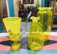 Photo showing all four canary yellow glass items on patterned table: Murano bird figurine, Blenko decanter, textured tumbler, and large vase