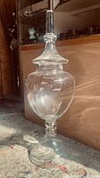 Full view of large clear glass apothecary jar with lid standing on carpeted floor, showing height and shape