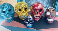 Front view of five colorful hand-painted Mexican folk art sugar skulls in blue, yellow, red, small blue, and brown, showing detailed floral and ornamental design work.