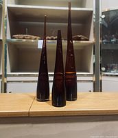 Front view of three dark amber long tapered decorative vases of varying heights on display shelf.