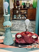 Both lamps together showing relative scale, color, and form. Blue lamp tall and vertical with gold accents. Red lamp shorter with flowing sculptural cutouts. Background shows vintage decor.