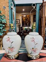 Front view of both matching vintage glass lamps showing cream bodies, pink floral painting, gold bases, and hardware for lamp fittings.