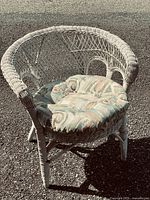 Vintage white wicker chair with curved back and a patterned padded cushion, shown outdoors on asphalt.