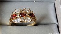 Front view of ring in display box showing three red stones and six clear stones set in gold-plated metal.