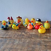 Photo shows 11 items: 10 rubber ducks in various costumes and one small plush bug toy, arranged in front of a plain wall on a wooden surface.