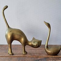Brass cat and swan figurines placed side by side on a wooden surface with a plain background.