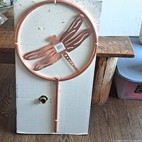 Front view of the circular copper frame with dragonfly design made of metal, attached copper tubing, and part of metal stake base on cardboard backing.