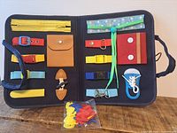 Open tote showing interior features like zipper pocket, various colored straps and buckles, laces, buttons, and small pouch for child skill development.