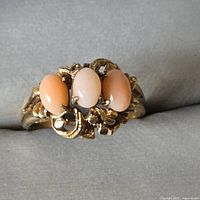 Front view of a gold-tone cocktail ring with three peach-colored oval cabochon stones set horizontally with decorative gold metal curling accents.